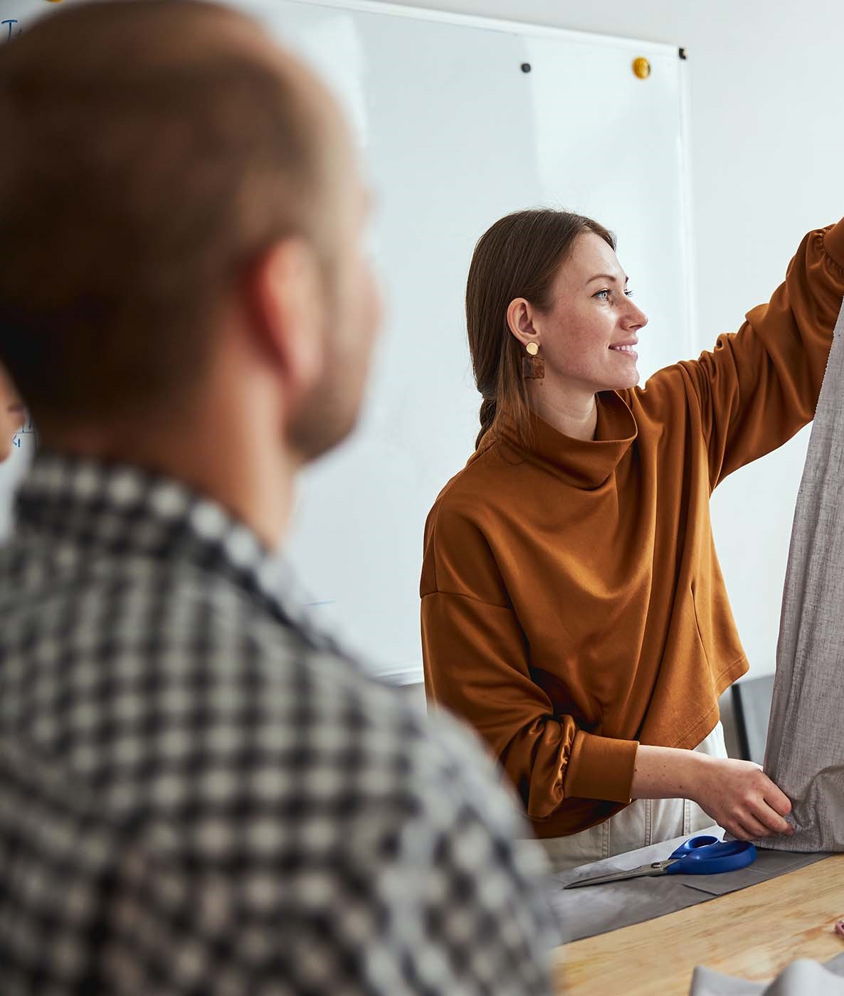 A woman in a brown sweater pointing at a whiteboard while a man in a checkered shirt looks on