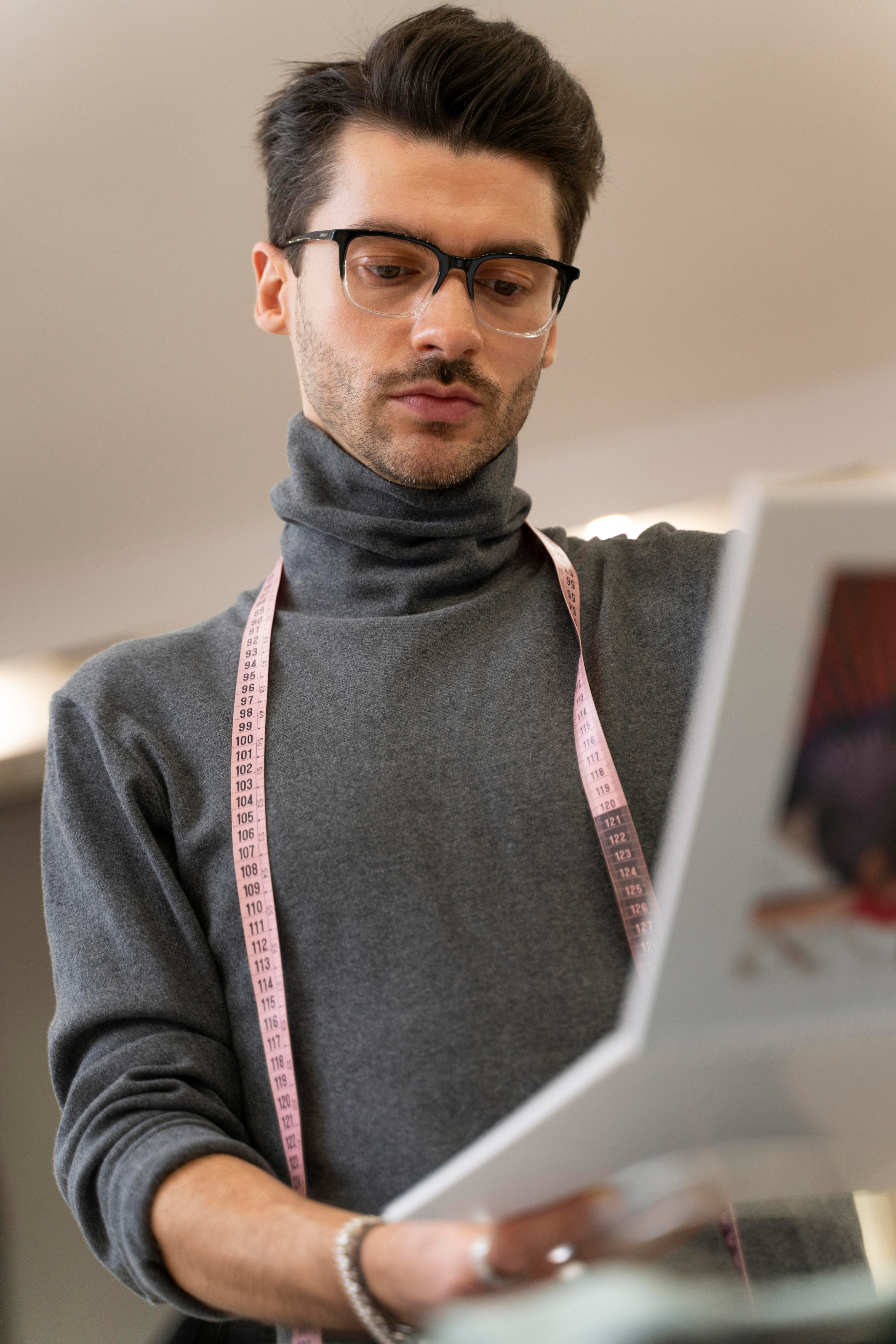 A man wearing glasses and a grey turtleneck with a pink measuring tape around his neck, looking at a document